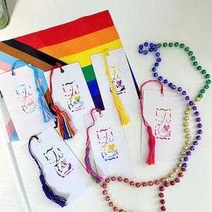 Puede incluir: Cinco marcapáginas rectangulares de papel blanco, cada uno con un diseño abstracto colorido único y una borla de hilo a juego. Los marcapáginas están dispuestos sobre una bandera arcoíris, acompañados por un largo collar de cuentas verdes, moradas, doradas y rojas.