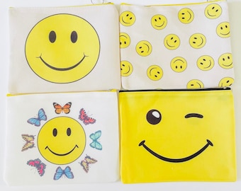 May include: Set of four yellow canvas zipper pouches with smiley face designs. One pouch features a single large smiley face, another has a smiley face with butterflies, another has a winking smiley face, and the last has a repeating pattern of small smiley faces.