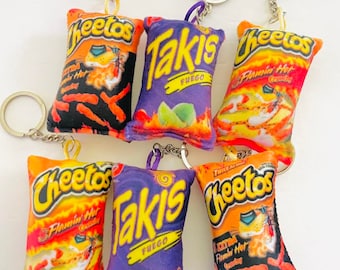 May include: Six plush keychain bags shaped like bags of popular snack chips. The bags are orange, purple, and black with the brand names "Cheetos" and "Takis" printed on them. The keychains are made of silver metal.