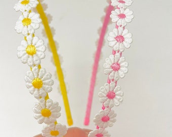 May include: Two floral headbands with white daisy-shaped embellishments. One headband is yellow and the other is pink.