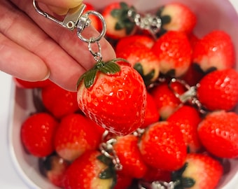 May include: A close-up of a red strawberry keychain with a silver metal clasp. The keychain is surrounded by other red strawberry keychains in a white bowl.