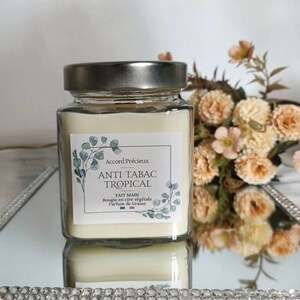 Peut inclure: Une bougie en verre carrée avec un couvercle argenté. L'étiquette indique "Accord Précieux Anti-Tabac Tropical Fait Main Bougie en cire végétale Parfum de Grasse".