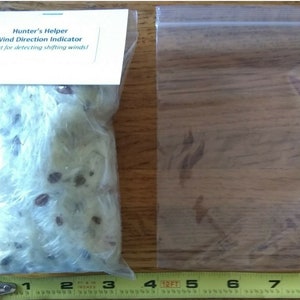 May include: A clear plastic bag containing white fluffy material and small brown seeds. The bag is labeled "Hunter's Helper Wind Direction Indicator Great for detecting shifting winds!"