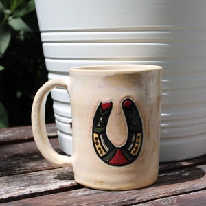 May include: Cream-colored ceramic mug with a horseshoe design. The horseshoe is black with red, green, and gold accents. The mug has a curved handle and sits on a wooden surface.