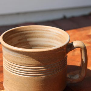 Puede incluir: Taza de cerámica beige con asa, de aspecto rústico y hecha a mano. La taza presenta líneas horizontales en el cuerpo. El interior es de color beige claro. Un artículo de cocina artesanal, ideal para café o té.