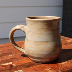 Puede incluir: Taza de cerámica hecha a mano en tonos beige y marrón claro. La taza tiene un asa grande y una abertura amplia. El exterior presenta bandas horizontales de diferentes tonos, lo que le da un aspecto rústico. Ideal para café o té.