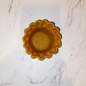 Puede incluir: Un pequeño cuenco decorativo amarillo con un borde festoneado. El cuenco está hecho de un material liso y brillante y tiene un borde ligeramente elevado.