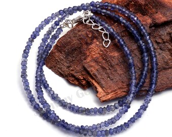 May include: A strand of blue gemstone beads with a silver clasp. The beads are faceted and have a slightly irregular shape. The necklace is displayed on a piece of wood.