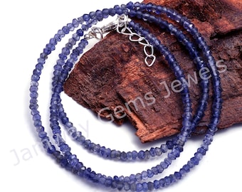 May include: A double strand necklace made of small, faceted, blue gemstone beads. The necklace has a silver clasp and is displayed on a piece of wood.