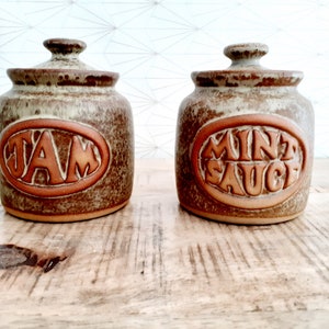 Op de afbeelding: Twee keramische potten met deksels, een met het label "JAM" en de andere met "MINT SAUCE". De potten zijn bruin en groen met een gespikkeld glazuur.