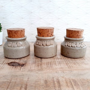 Może przedstawiać: Trzy ceramiczne słoiki na przyprawy z korkowymi pokrywkami. Słoiki są oznaczone etykietami "Dill", "Thyme" i "Parsley".