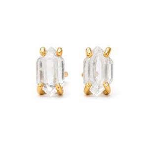 Peut inclure: Une paire de boucles d'oreilles à clous avec des cristaux hexagonaux allongés et transparents montés sur des griffes en métal doré. Chaque cristal présente des pointes aux extrémités, maintenu par quatre petites griffes métalliques.