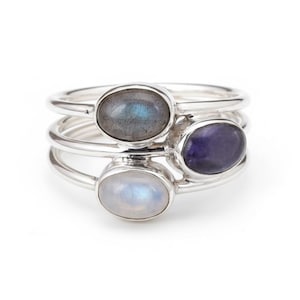 Pode incluir: Um conjunto de anéis empilháveis com três bandas finas de prata, cada uma com uma pedra preciosa oval. As pedras incluem uma labradorite cinzenta com iridescência azul, uma ametista roxa profunda e uma pedra da lua branca translúcida.