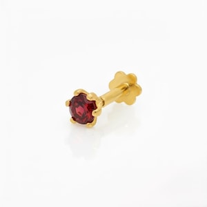 May include: A gold stud earring featuring a round, faceted deep red gemstone held in a six-prong setting. The post is attached to a flat, flower-shaped backing, providing a secure fit for this piece of jewelry designed for ear piercings.