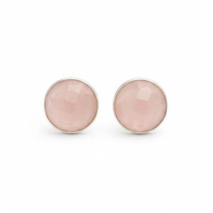 Puede incluir: Un par de pendientes de botón redondos con piedras de color rosa pálido facetadas engastadas en un bisel de metal plateado liso. Las gemas circulares tienen un acabado pulido que refleja la luz, creando un sutil patrón geométrico en la superficie.