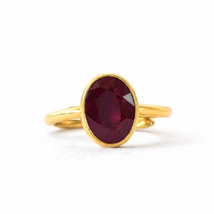 May include: A gold ring featuring a large, oval-cut, deep red gemstone set in a smooth bezel setting. The band of the ring is thin, rounded, and polished, showcasing a bright yellow gold tone that contrasts with the dark red of the central stone.