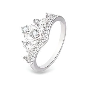 Puede incluir: Un anillo de corona plateado con piedras preciosas transparentes. El anillo presenta un diseño de corona con elementos en forma de corazón y redondeados, incrustado con pequeñas piedras brillantes. La banda es lisa y pulida.