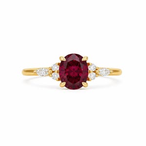 May include: A gold ring featuring a central oval-cut deep red gemstone held by four prongs. The band is accented on each side by two small round-cut clear stones and one marquise-cut clear stone, creating a symmetrical cluster design.