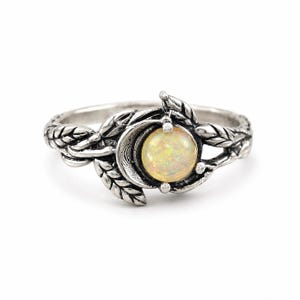 May include: A silver-toned ring featuring a central round iridescent opal stone set within a crescent moon shape. The band is crafted with a detailed relief pattern of overlapping leaves that extend along the sides of the ring, creating a textured finish.