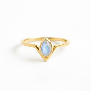 Könnte beinhalten: Ein goldfarbener Ring mit einem tropfenförmigen, irisierenden Edelstein. Das Band des Rings hat ein zartes, V-förmiges Design, und der Edelstein zeigt Schattierungen von Blau und Weiß. Der Ring ist vor einem weißen Hintergrund platziert.
