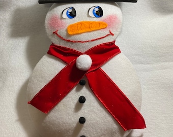 May include: A white felt snowman ornament with a black felt hat, red felt scarf, and black felt buttons. The snowman has a carrot nose, blue eyes, and rosy cheeks.