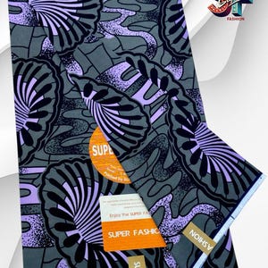 May include: A folded piece of fabric featuring a dark gray background with a repeating pattern of black and light purple fan-shaped motifs. The textile includes a circular orange sticker and a rectangular white label with the text Super Fashion.