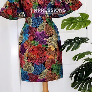 Könnte beinhalten: Ein farbenfrohes Kleid mit einem Blatt-Muster auf schwarzem Hintergrund. Das Kleid hat einen Rüschenkragen und kurze Ärmel. Das Kleid befindet sich auf einer Holzpuppe.