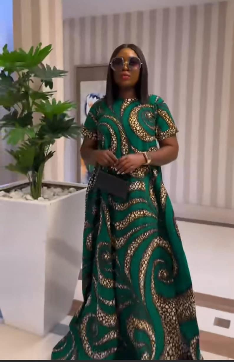 May include: A floor-length dress featuring a vibrant green base with a repeating pattern of swirling, textured gold and black circular motifs. The garment has short sleeves, a round neckline, and a loose, flowing silhouette that reaches the floor.