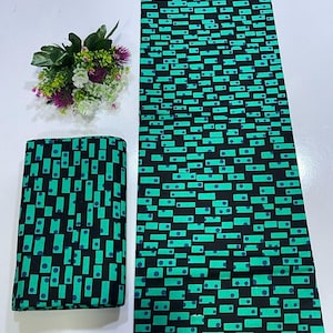 May include: A length of fabric and a folded square of textile featuring a repeating geometric pattern of black rectangles and dots on a vibrant teal background. The fabric displays a consistent print of small dark blue circles centered within rectangular shapes.