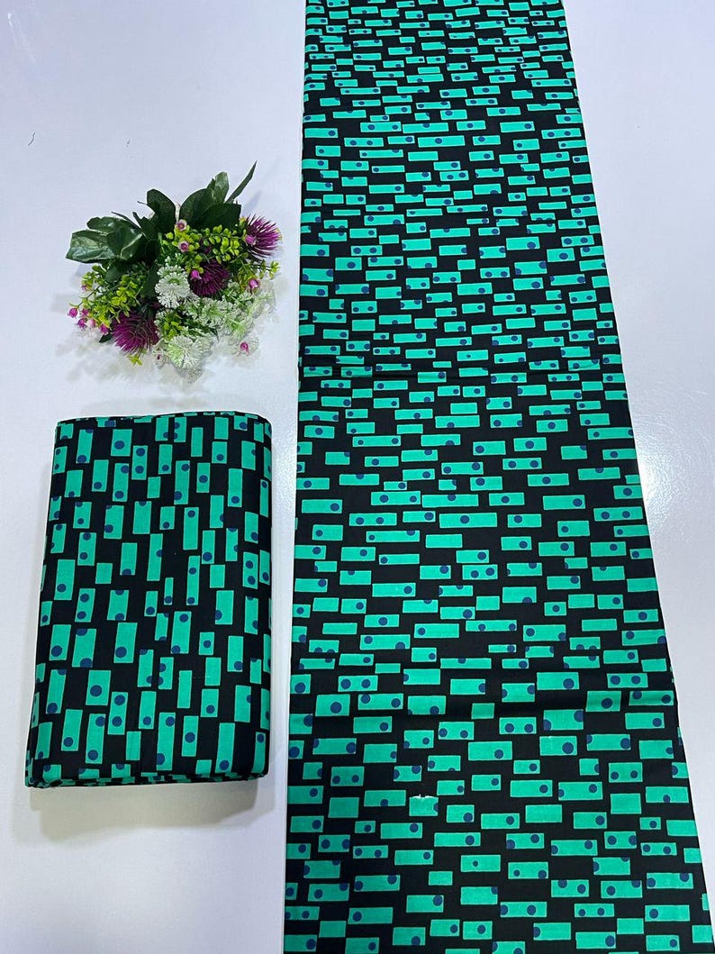 May include: A length of fabric and a folded square of textile featuring a repeating geometric pattern of black rectangles and dots on a vibrant teal background. The fabric displays a consistent print of small dark blue circles centered within rectangular shapes.
