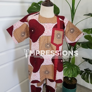 Könnte beinhalten: Ein zweiteiliges Outfit für ein Kind, bestehend aus einem kurzärmeligen Hemd und einer Hose. Der Stoff hat einen weißen Hintergrund mit einem roten und braunen geometrischen Muster. Das Hemd hat einen roten Saum entlang des Ausschnitts und eine rote Tasche auf der linken Seite. Die Hose hat einen roten Saum entlang des Bunds. Das Outfit wird auf einer Puppe präsentiert.