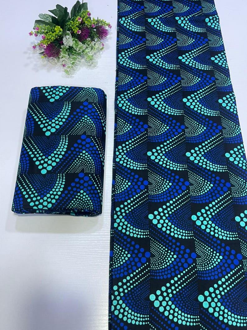 May include: A folded piece of fabric and a long strip of the same material, both featuring a black background with a repeating geometric pattern of blue and turquoise dots arranged in chevron-like waves, accompanied by a small floral bouquet.
