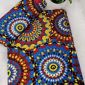 May include: A folded piece of fabric featuring a vibrant, repeating circular mandala pattern in shades of red, yellow, blue, and black. The textile displays intricate geometric details and includes the printed text HITARGET 3071302 along the edge.
