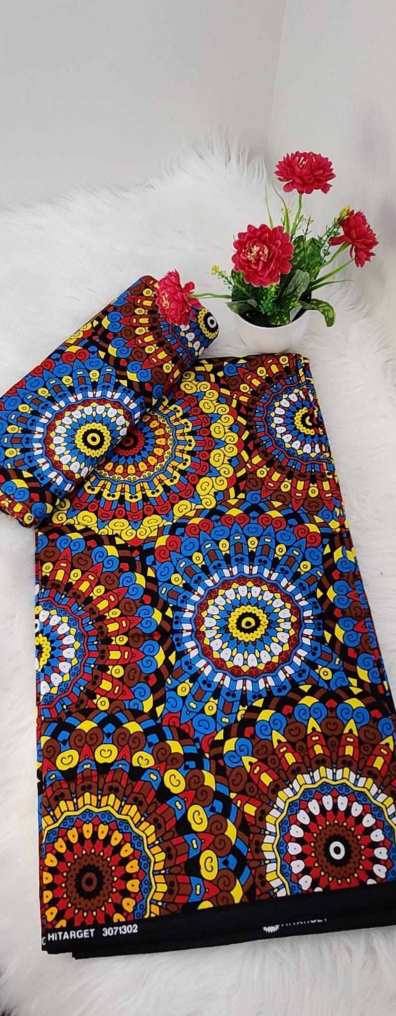May include: A folded piece of fabric featuring a vibrant, repeating circular mandala pattern in shades of red, yellow, blue, and black. The textile displays intricate geometric details and includes the printed text HITARGET 3071302 along the edge.
