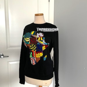 Peut inclure: Sweat-shirt noir avec un graphique coloré du continent africain. Le graphique est composé de différents tissus à motifs. Le sweat-shirt porte le texte "VMPRESSIONS" imprimé sur le devant.