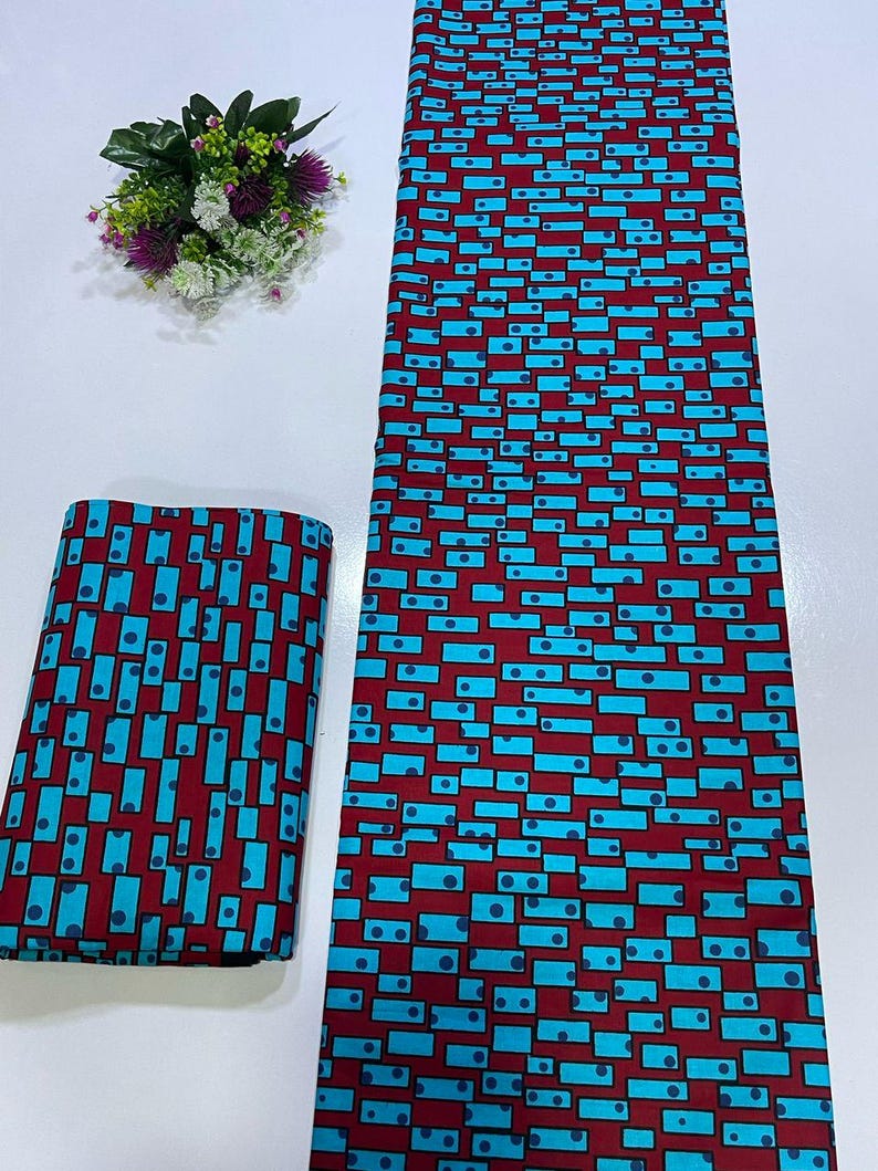 May include: A length of fabric and a folded square of the same material, both featuring a repeating geometric pattern of bright cyan rectangles and small dark blue circles set against a deep maroon background, accompanied by a small floral bouquet.