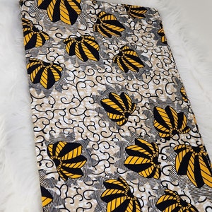 May include: A folded piece of fabric featuring a repeating pattern of yellow and black fan-shaped motifs on a beige background with black swirling lines. A small rectangular tag with the text "EDITION" is attached to the bottom right corner.