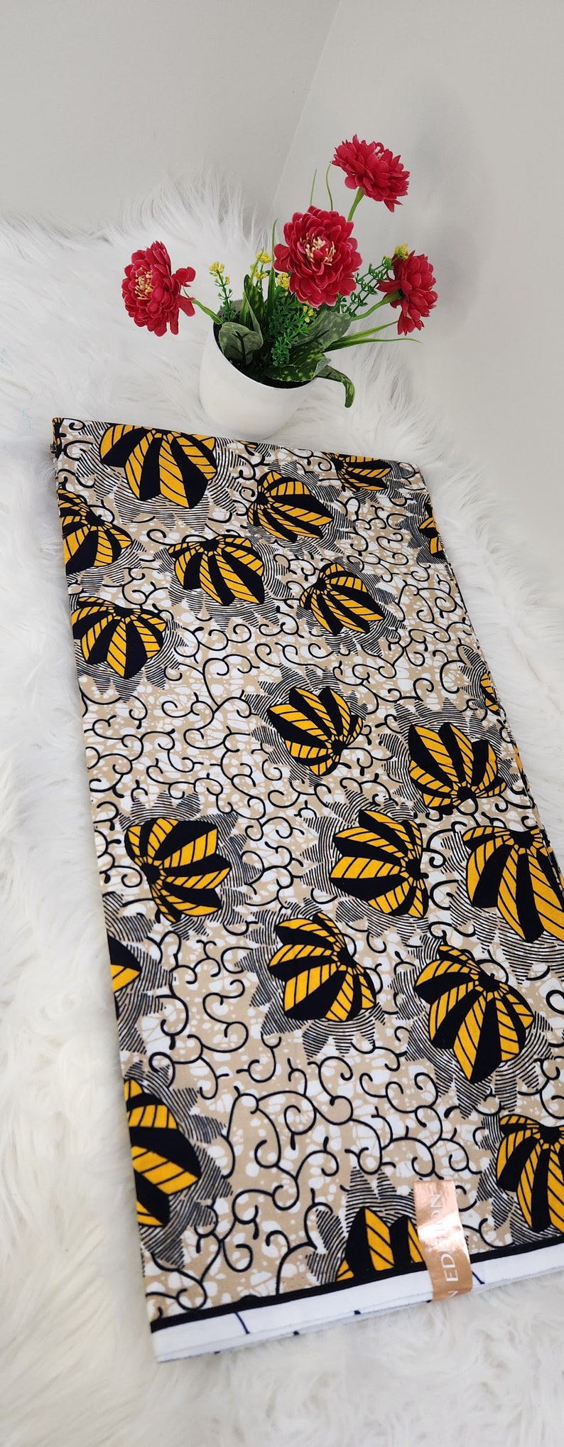 May include: A folded piece of fabric featuring a repeating pattern of yellow and black fan-shaped motifs on a beige background with black swirling lines. A small rectangular tag with the text "EDITION" is attached to the bottom right corner.