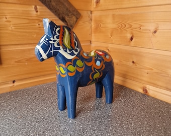 May include: A blue wooden Dala horse with traditional Swedish folk art painted on it. The horse has a white mane and tail, and the design features red, yellow, green, and blue colours.