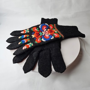 May include: A pair of black knitted gloves with colorful embroidered floral designs. The gloves are folded together and displayed on a white surface.