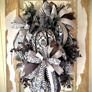 Puede incluir: Una corona decorativa de calabaza con una calabaza con estampado de damasco en el centro, rodeada de cintas negras, blancas y grises. La corona incluye varias texturas y patrones, creando un diseño en capas para la decoración de temporada.