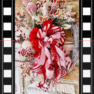 Op de afbeelding: Een rode en witte kerstkrans met snoepstokken, een rood-wit gestreepte strik, peperkoekjes en witte sneeuwvlokken. De krans is op een witgeschilderde houten achtergrond met de tekst "Booker's Finest".