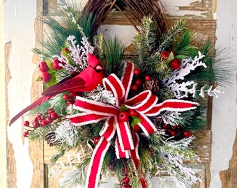 May include: A red cardinal bird perched on a winter wreath with red and white striped ribbon, pine branches, red berries, and frosted pine needles.
