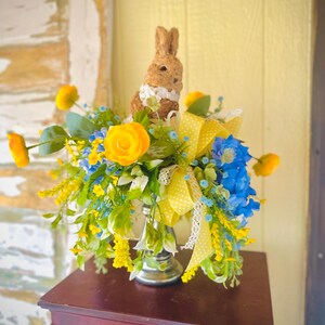 May include: A decorative Easter arrangement featuring a brown bunny figurine with a white collar, surrounded by yellow and blue artificial flowers. The arrangement includes a yellow polka dot ribbon and is displayed in a silver vase on a dark wooden surface.
