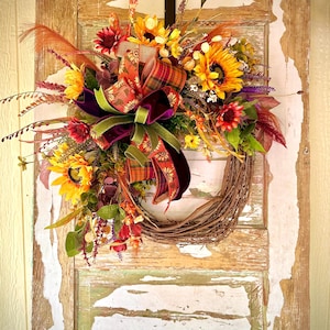 Puede incluir: Una corona de otoño con girasoles, flores rojas y amarillas y un gran lazo a cuadros. La corona está hecha de ramitas tejidas y cuelga de una puerta de madera desgastada. El lazo tiene una cinta de terciopelo y detalles verdes.