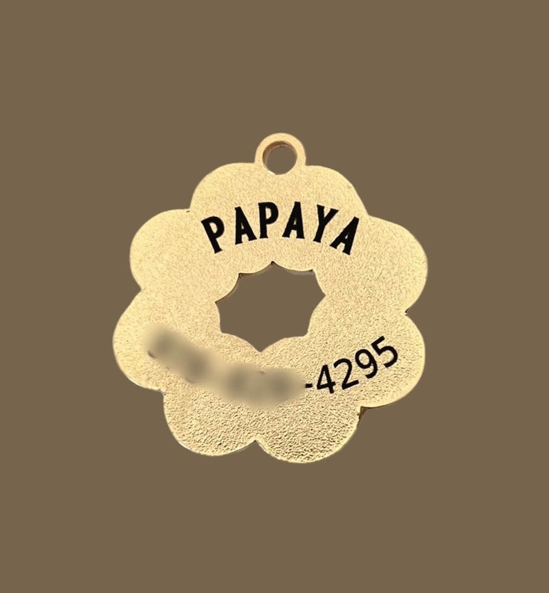 K&ouml;nnte beinhalten: Goldfarbener, blumenf&ouml;rmiger Anh&auml;nger aus Metall mit dem Wort "PAPAYA" und der Nummer "-4295", die in Schwarz eingraviert sind.
