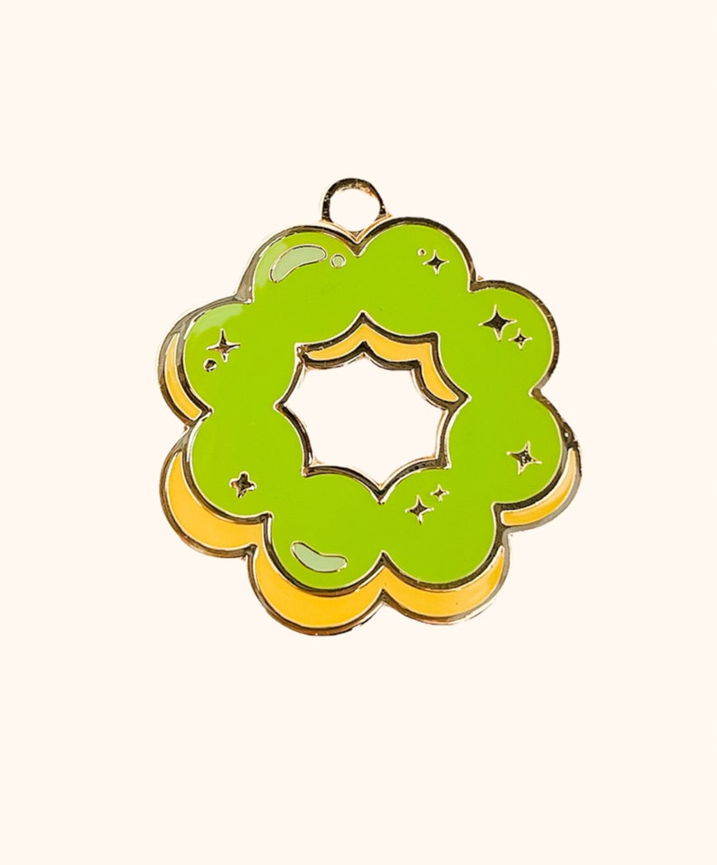 K&ouml;nnte beinhalten: Eine gr&uuml;ne und gelbe Emaille-Anstecknadel in Form eines Donuts mit einem sternf&ouml;rmigen Loch in der Mitte. Der Donut hat eine gl&auml;nzende Oberfl&auml;che und ist mit kleinen wei&szlig;en Sternen verziert.