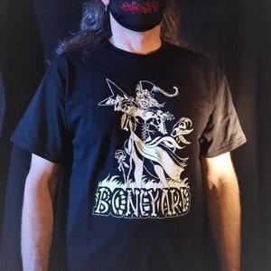 Peut inclure: T-shirt noir avec un motif blanc d'un squelette jouant de la guitare, le mot "BONEYARD" en dessous. La personne porte un masque noir avec du texte rouge, une casquette noire et des lunettes de soleil.