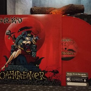 Peut inclure: Un disque vinyle rouge avec une étiquette noire comportant le texte "Boneyard Cashbreaker" et une illustration en noir et blanc d'un corbeau. Le disque est sur un fond rouge avec une illustration en noir et blanc d'une femme assise sur un trône.