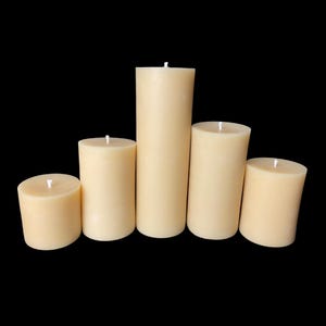 May include: Five unscented pillar candles in various sizes, all a pale yellow color, arranged in a row on a black background.